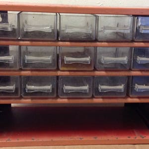 以下が含まれることがあります： 複数の引き出しが付いた金属製の収納キャビネット。キャビネットはオレンジ色に塗装されており、金属製のフレームがあります。引き出しは透明なプラスチック製で、小さなアイテムを収納するように設計されています。