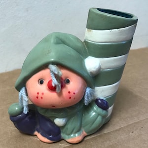 May include: A ceramic Christmas elf figurine with a green hat and a striped candy cane. The elf is holding a purple and green object in one hand and has a red nose and rosy cheeks.