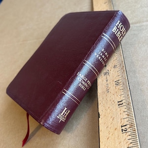 以下が含まれることがあります： 背表紙に金文字が刻まれたバーガンディ色のHoly Bible King James Version Large Print Compact聖書。木製の定規の横に置かれており、長さは約16.5cmであることが示されています。赤いリボンのしおりが付いています。