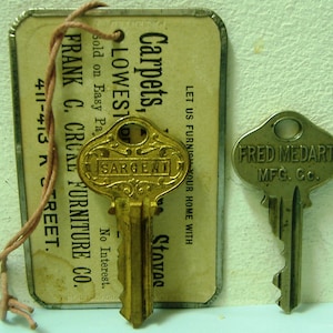 May include: A vintage key with a gold finish and ornate design, labeled "Sargent". The key is attached to a small, rectangular tag with the text "Carpets, LOWEST! Sold on Easy Pa. No Interest. FRANK C. CROAL FURNITURE CO. 411-413 N STREET."  The tag is attached to a string. A second, smaller key with a silver finish is labeled "FRED MEDART MFG. CO."