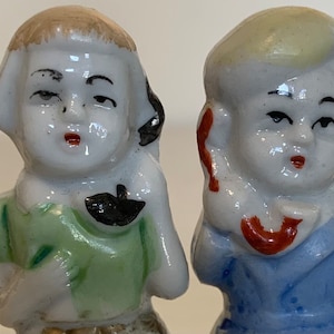 May include: Two porcelain figurines of children, one wearing a green shirt and the other wearing a blue shirt. Both figurines have painted facial features and hair.
