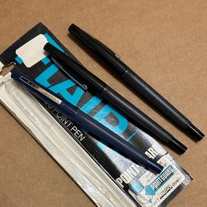 Puede incluir: Tres bolígrafos, dos negros y uno azul marino, se muestran sobre una superficie marrón. El bolígrafo azul marino aún está en su embalaje original, que tiene el texto "POINT PEN" y "POINT GUARD". El embalaje también tiene el precio de 59 ¢.