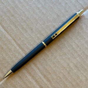 Op de afbeelding: Een zwarte pen met gouden details. De pen heeft een matte afwerking en een gouden clip en punt. Twee kleine hartvormige details zijn zichtbaar op de clip. De pen ligt op een kartonnen oppervlak.