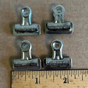 May include: Four vintage silver-coloured Boston Clip No. 0 paper clips. Each clip has the text "BOSTON CLIP NO. 0 STATESVILLE, N.C." stamped on the front. A wooden ruler is at the bottom of the image.