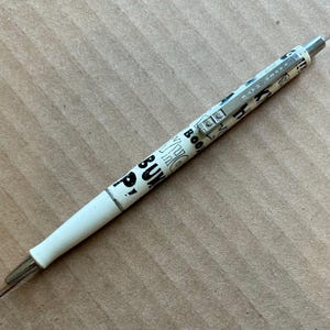May include: A white retractable ballpoint pen featuring a silver metal clip and a barrel decorated with black printed text including the words WHO, BUM, and BOOM. The pen has a white plastic grip section and a silver metal tip and end cap.