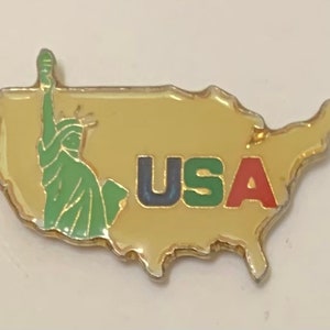 May include: A gold-colored pin in the shape of the United States, featuring the Statue of Liberty in green and the word "USA" in blue, green, and red. The pin is a souvenir or collectible item.