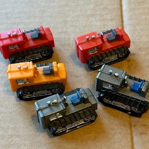 May include: Five toy construction vehicles in red, orange, and grey. Each miniature bulldozer has black treads and a detailed cab. The toys are arranged on a light brown surface.