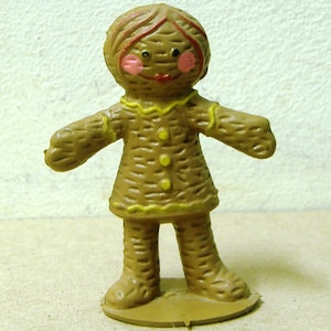 May include: A brown gingerbread person figurine with yellow icing details and a painted face. The figurine has a red mouth, pink cheeks, and black eyes.