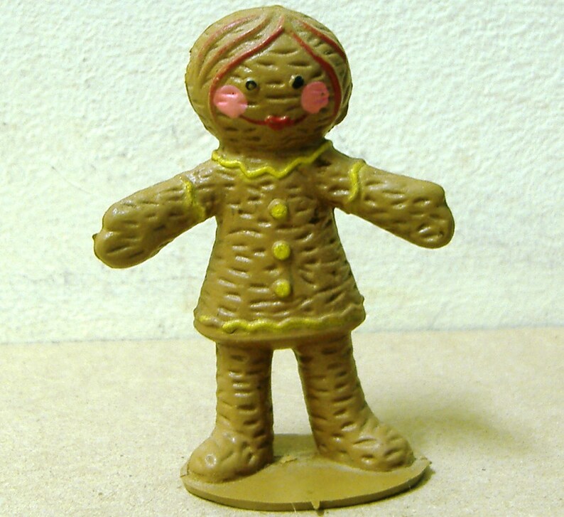 May include: A brown gingerbread person figurine with yellow icing details and a painted face. The figurine has a red mouth, pink cheeks, and black eyes.