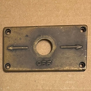 May include: A brass switch plate with two arrows pointing in opposite directions and the word "OFF" inscribed below a round hole.