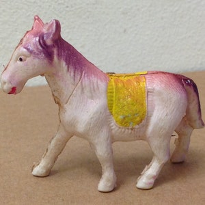 Könnte beinhalten: Ein weißes Plastik-Spielzeugpferd mit einer lila Mähne und einem lila Schwanz. Das Pferd hat einen gelben Sattel und steht auf einer braunen Oberfläche.