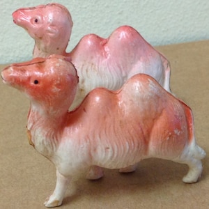 Könnte beinhalten: Zwei rosafarbene und weiße Keramik-Kamelfiguren. Die Kamele stehen nebeneinander, das Kamel links schaut nach rechts und das Kamel rechts schaut nach links.