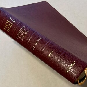 Könnte beinhalten: Eine burgunderrote, ledergebundene Heilige Bibel mit goldener Schrift. Das Buch ist ein Scofield Study System, NIV, herausgegeben von Oxford. Ein goldenes Leseband ist sichtbar. Der Einband ist strukturiert.