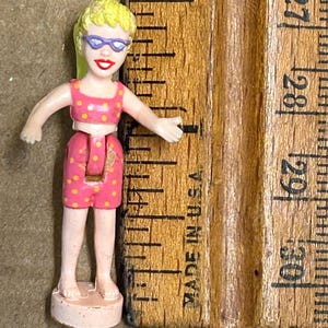 May include: A small plastic doll wearing a pink and white polka dot swimsuit and blue sunglasses. The doll is standing on a pink base and is about 2 inches tall.