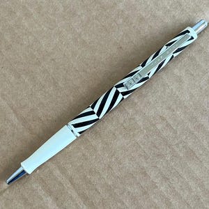 May include: A black and white patterned ballpoint pen with a silver clip. The pen has a white tip and a black and white chevron design on the barrel. The pen is lying on a brown surface.