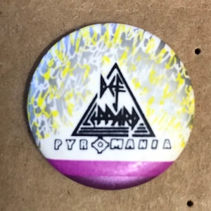 Könnte beinhalten: Ein weißer Knopf mit einem schwarzen Dreieck und dem Text "Def Leppard" im Dreieck. Der Text "Pyromania" befindet sich unter dem Dreieck. Der Knopf hat ein gelbes und graues Wirbelmuster um das Dreieck und einen violetten Rand.
