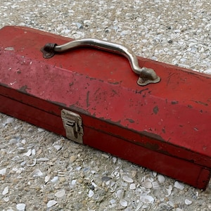 May include: A red metal toolbox with a silver handle and a latch. The toolbox is slightly weathered and shows signs of use.