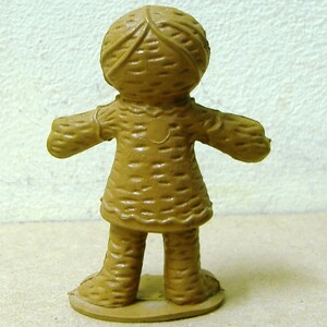 May include: A brown plastic doll with a woven pattern on its clothing and head. The doll has a round base and is standing with its arms outstretched.