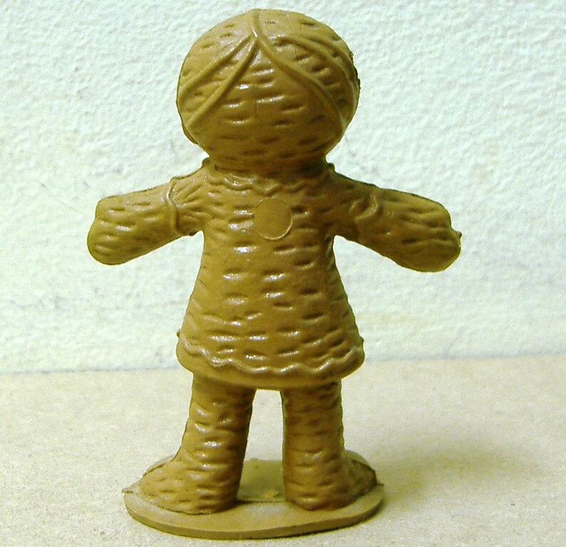 May include: A brown plastic doll with a woven pattern on its clothing and head. The doll has a round base and is standing with its arms outstretched.