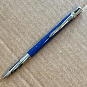 Op de afbeelding: Een blauwe pen met zilveren details en een gespikkelde afwerking. De pen heeft een zilveren clip en punt, en een zilveren bovenkant met een zwart ontwerp. De pen ligt op een bruin oppervlak.