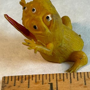 Puede incluir: Figura de rana amarilla con múltiples ojos y una lengua roja extendida. La rana está colocada sobre una superficie blanca, con una regla de madera que muestra medidas en pulgadas en primer plano. La figura tiene una superficie texturizada.