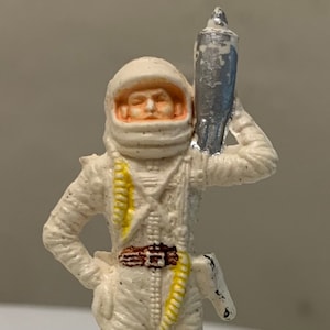 Puede incluir: Una figura de juguete de astronauta de plástico blanco que lleva un casco y sostiene un cohete plateado. El astronauta tiene un cinturón marrón y correas amarillas en el traje.