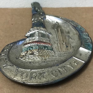 May include: A silver metal ashtray featuring a raised image of the Statue of Liberty and the Empire State Building. The ashtray is shaped like a crescent moon and has the words "NEW YORK CITY" around the edge.