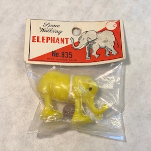 May include: A yellow plastic toy elephant with a white string around its body. The elephant is in a clear plastic bag with a red and white card that reads "Space Walking Elephant No. 835 Made in Hong Kong".