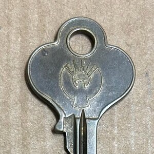 Puede incluir: Una llave de latón vintage con una cabeza en forma de trébol. La llave tiene un agujero redondo en la cabeza y la palabra "EAGLE" está grabada en la cabeza.