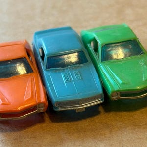 Könnte beinhalten: Drei Spielzeugautos in Orange, Blau und Grün. Die Autos sind kleine Plastikmodelle mit klaren Windschutzscheiben und silberfarbenen Zierleisten. Das orangefarbene Auto befindet sich links, das blaue in der Mitte und das grüne rechts.