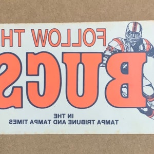 Könnte beinhalten: Ein weißer und orangefarbener Vintage-Aufkleber mit dem Text "FOLLOW THE BUGS" in orangefarbenen Buchstaben mit schwarzem Umriss. Der Text "TAMPA TRIBUNE AND THE TIMES" ist in Schwarz am unteren Rand des Aufklebers gedruckt. Eine Comic-Illustration eines Footballspielers in weiß-orangefarbener Uniform rennt nach rechts.