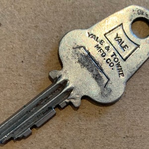 以下が含まれることがあります： 銀色の Yale & Towne Mfg. Co. の鍵。鍵穴は上部にあり、鍵の端はギザギザになっている。鍵は少しすり減っている。