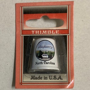 Puede incluir: Dedal de cerámica blanca con una imagen de un coche de policía y las palabras "Mayberry, Carolina del Norte". El dedal está empaquetado en una caja de cartón roja y blanca con la palabra "THIMBLE" impresa en ella.