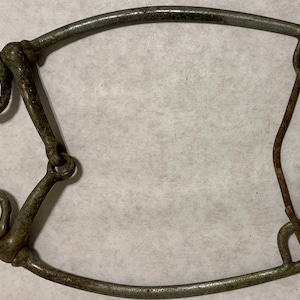 Puede incluir: Un antiguo bocado de caballo hecho de metal desgastado. El bocado presenta una embocadura curva, dos anillas y una barra de conexión. El metal tiene un aspecto desgastado, lo que sugiere su uso. El diseño es típico del equipo ecuestre.