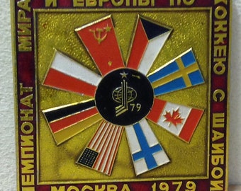 Puede incluir: Un pin de metal dorado con un borde rojo. El centro presenta un disco negro con un palo de hockey estilizado y el año 1979. Alrededor del disco se encuentran banderas de diferentes países, incluyendo la URSS, EE. UU., Canadá y Suecia. El texto "ЧЕМПИОНАТ МИРА И ЕВРОПЫ ПО ХОККЕЮ С ШАЙБОЙ" está escrito alrededor del borde. El texto "МОСКВА 1979" está en la parte inferior.
