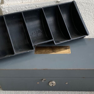 Peut inclure: Une boîte à monnaie en métal gris avec une serrure et un plateau en plastique noir à cinq compartiments. Le plateau porte l'inscription "ASCO".