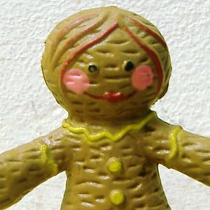 May include: A gingerbread person figurine with a brown body, yellow trim, and pink cheeks. The figurine has a red mouth and brown hair.
