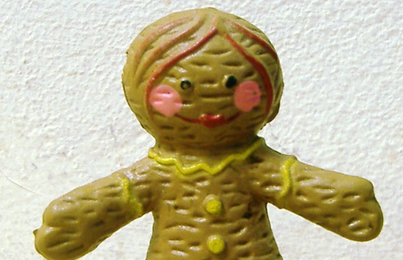 May include: A gingerbread person figurine with a brown body, yellow trim, and pink cheeks. The figurine has a red mouth and brown hair.