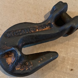 Könnte beinhalten: Ein robuster, schwarzer Metallhaken mit dem Text "WEISSALLOY CO." auf der Seite. Der Haken hat ein gebogenes Design mit einer breiten Öffnung und einer Verbindungsschlaufe. Das Metall weist Gebrauchsspuren und Rost auf.