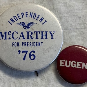 May include: Two vintage political campaign buttons. The larger button is white with blue text and an eagle graphic. It reads "Independent McCarthy for President '76". The smaller button is maroon with white text that reads "Eugene".