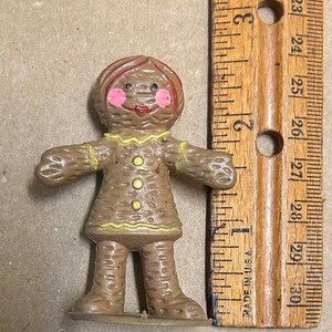 May include: A brown gingerbread person figurine with a red hair, pink cheeks, and yellow buttons. The figurine is about 3 inches tall and is standing on a white base.