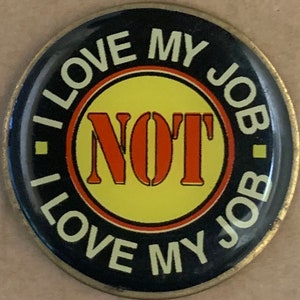 Op de afbeelding: Een ronde zwarte en gouden knop met de tekst "I LOVE MY JOB - NOT I LOVE MY JOB" in witte letters. De woorden "NOT" staan in rode en oranje letters op een gele cirkel.