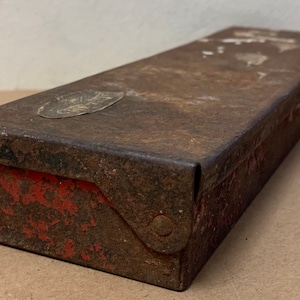 Peut inclure: Une boîte en métal rouillée avec un intérieur rouge et un autocollant argenté sur le dessus. La boîte a un couvercle à charnières et un fermoir en métal.