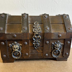 May include: A small, wooden treasure chest with a dark brown finish. The chest features metal accents, including corner brackets, a decorative clasp, and lion's head handles. The chest is rectangular and has a hinged lid.