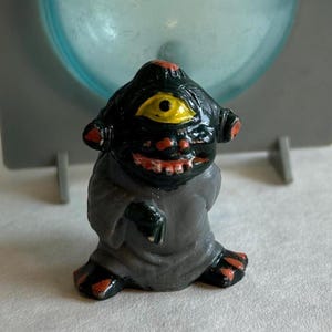 May include: A small, black figurine of a one-eyed creature with a yellow iris, red accents, and a gray robe. The figurine has a wide, toothy grin and is posed in a seated position. The background includes a light blue and gray structure.