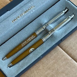 May include: Two pens in a blue case. The pens have a silver top and a brown barrel with the text "PAPER COMPANY Continuous Fair Share". The case is labeled "PAPER MATE".