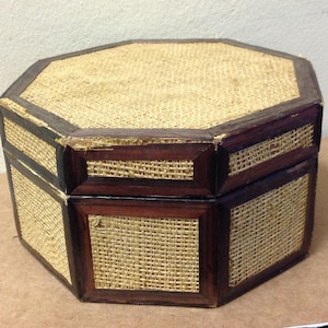 Op de afbeelding: Achtekantige houten doos met een geweven strooien deksel en zijkanten. De doos heeft een donkerbruine houten rand en is versierd met een geweven strooien patroon.