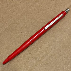 May include: A red ballpoint pen with a silver clip and tip. The pen is a vibrant red color and has a sleek, streamlined design. The silver clip has the brand name on it. The pen is lying on a brown surface.