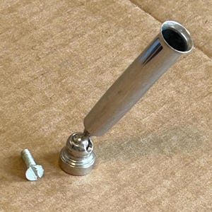 May include: A silver-toned pen holder with a wooden handle and a small screw. The pen holder has a rounded base and a cylindrical opening for a pen. The screw is silver and has a slotted head.