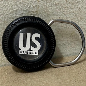 Könnte beinhalten: Ein schwarzer Gummi-Flaschenverschluss mit einem silbernen Metallgriff. Der Verschluss hat die weißen Buchstaben "US" über dem Wort "RUBBER" in einem weißen Rechteck. Der Verschluss ist rund mit einem geriffelten Rand.